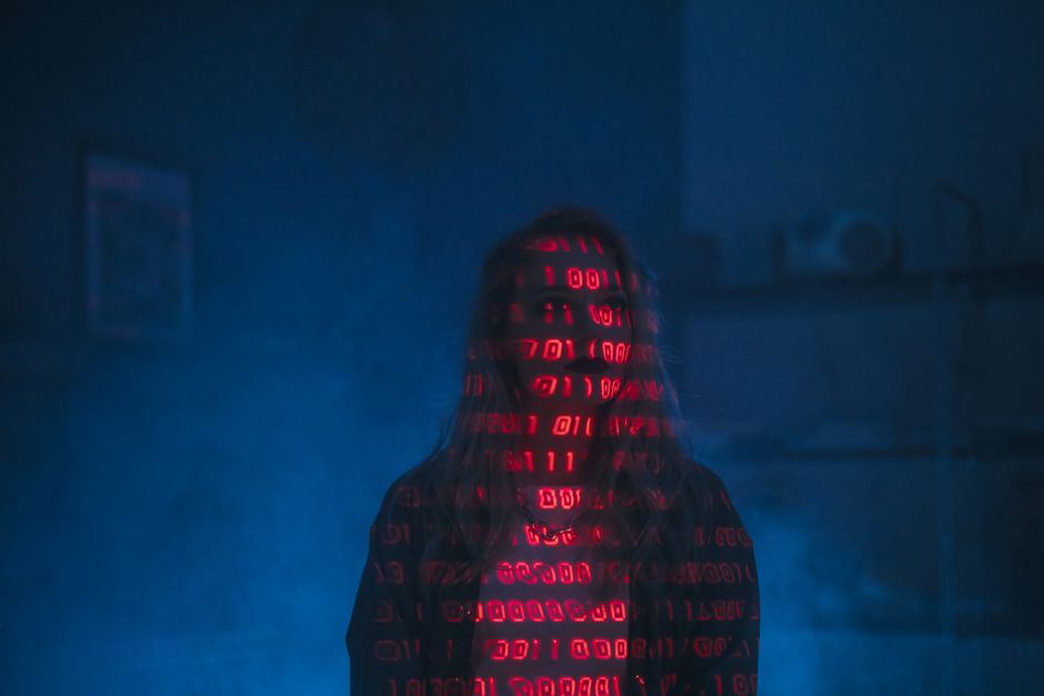A person standing in a dimly lit room with a dark background, their face partially obscured by long hair, illuminated by the red glow of ASCII-style digital code projecting onto their face and upper b